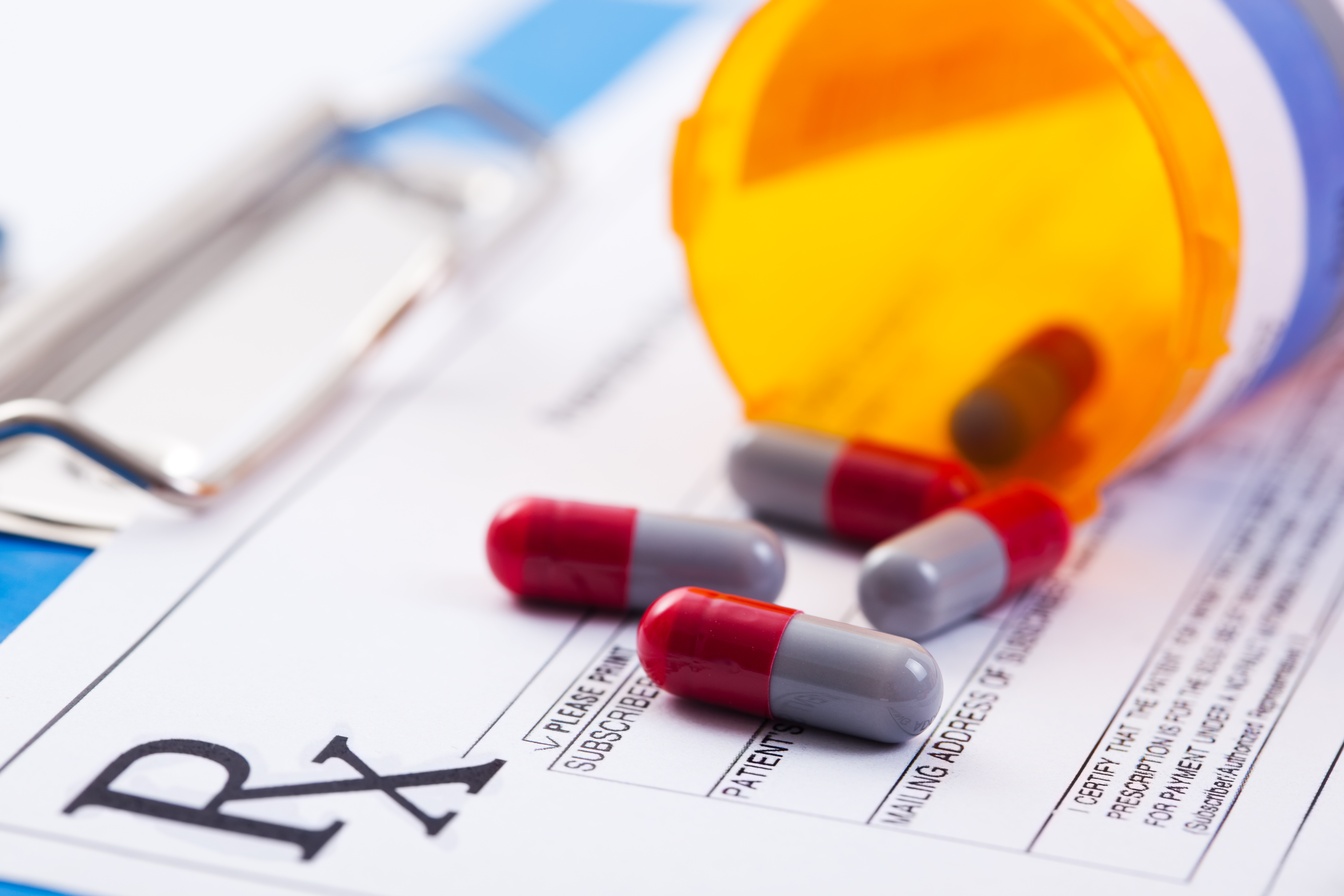 Red and blue pills spilling out of orange prescription bottle on top of prescription pad.